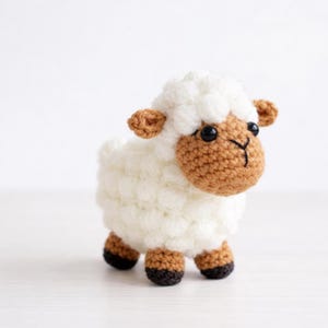 May include: A crocheted sheep toy with a fluffy white body, a brown face, ears, and feet, and black eyes. The sheep is standing on a white surface. This handmade soft toy is ideal for nursery decor or as a child's toy.