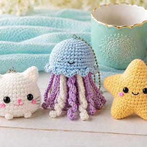 May include: Three crocheted keyrings: a white cat with pink details, a blue jellyfish with purple and white tentacles, and a yellow starfish. Each has a smiling face and a metal chain. A teal mug and small potted plants are in the background.