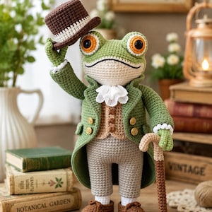 Könnte beinhalten: Eine gehäkelte Froschpuppe in einem grünen Gehrock, einer beigen Weste und einer hellgrauen Hose. Der Frosch hat orangefarbene Augen, einen weißen Rüschenkragen und braune Schuhe. Er hält einen braunen Zylinder in der Hand und einen Gehstock.