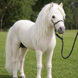 以下が含まれることがあります： クリームホワイトの糸で手編みされたかぎ針編みの馬のフィギュア。長く流れるような白いたてがみと尻尾が特徴です。馬には灰色の鼻口部と蹄があり、黒い手綱と頭部に付けられた細い黒いリードロープが装備されています。
