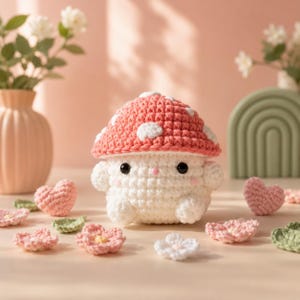 May include: A crocheted mushroom figure with a red and white spotted cap and a white body. The figure has black eyes and a small pink nose. Scattered around the mushroom are crocheted hearts, flowers, and leaves in shades of pink and green.