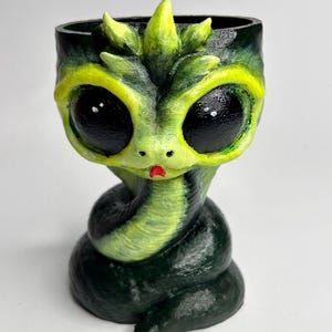 Könnte beinhalten: Ein skurriler, handbemalter Pflanztopf in Form eines Zeichentrickdrachen. Der Pflanztopf hat große, schwarze Augen, ein grün-gelbes Farbschema und eine gewundene, schlangenartige Basis. Das Gesicht des Drachen hat einen kleinen roten Mund und eine Krone aus grünen Blättern.