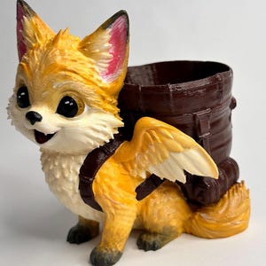 May include: A whimsical, hand-painted figurine of a fox with large eyes, wings, and a brown barrel on its back. The fox is primarily yellow and white with pink accents on its ears. The barrel is dark brown.