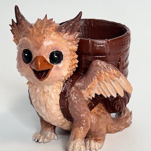 May include: A whimsical, hand-painted griffin planter. The sculpture features a smiling griffin with large black eyes, tan and brown feathers, and a brown barrel-shaped planter on its back. The planter is designed for small plants or decorative items.