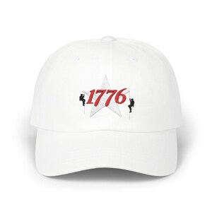 Könnte beinhalten: Weiße Baseballkappe mit gebogenem Schirm. Die Kappe zeigt ein Stern-Design mit der Zahl "1776" in roter Schrift und zwei kleinen Figuren, die an Seilen auf beiden Seiten des Sterns klettern.