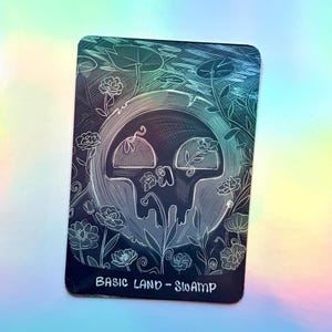 Puede incluir: Una tarjeta coleccionable rectangular con una ilustración de líneas blancas de una calavera centrada en una forma de luna circular, rodeada de flores y nenúfares. El texto "BASIC LAND - SWAMP" está impreso en blanco en la parte inferior.