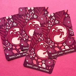 May include: Three rectangular playing cards featuring a deep magenta and pink floral pattern with a central circular mountain icon. Each card displays the text BASIC LAND - MOUNTAIN at the bottom in white capital letters against a dark purple border.
