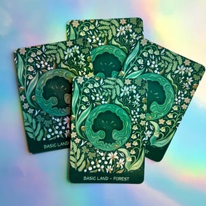 May include: Four rectangular game cards featuring a dark green background with a central circular emblem of a stylized tree. The surrounding area is decorated with detailed illustrations of white flowers, leaves, and ferns. Each card displays the text BASIC LAND - FOREST.