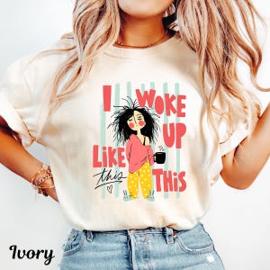 May include: An ivory short-sleeve t-shirt featuring a graphic of a person with messy hair, a pink top, and yellow polka-dot pants holding a black mug. The text reads I WOKE UP LIKE THIS in red letters, with light blue vertical stripes in the background.