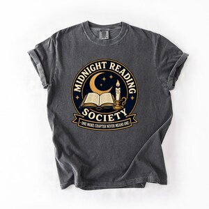 May include: A charcoal gray short-sleeve t-shirt featuring a circular graphic with a crescent moon, stars, an open book, and a lit candle. The text reads MIDNIGHT READING SOCIETY and ONE MORE CHAPTER NEVER MEANS ONE in a bold, stylized font.