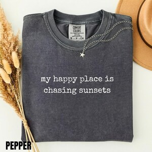 May include: A charcoal grey short-sleeve t-shirt with a vintage wash, featuring the white text "my happy place is chasing sunsets" printed on the chest. The shirt has a crew neckline and a Comfort Colors brand label inside the collar.