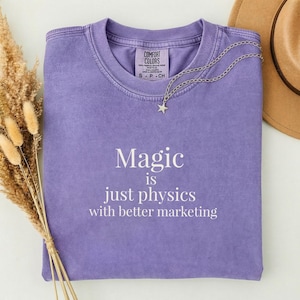 Puede incluir: Una camiseta morada de manga corta con cuello redondo y etiqueta Comfort Colors. La prenda presenta el texto impreso en blanco: Magic is just physics with better marketing. Un collar de cadena plateada con una pequeña estrella descansa sobre el cuello.