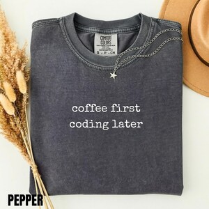 May include: A charcoal gray crew neck t-shirt with a vintage wash finish. The shirt features the white text "coffee first coding later" printed on the chest. A silver-toned chain necklace with a small star pendant rests on the collar.