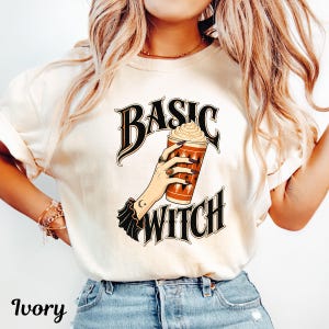 May include: An ivory t-shirt featuring a graphic of a hand holding a coffee cup with whipped cream. The text "BASIC WITCH" is printed in a stylised black font above and below the illustration. The hand has dark nail polish and rings.