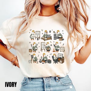 May include: An ivory t-shirt featuring a graphic print of cartoon ducks dressed as construction workers. The ducks are shown performing various tasks like planning, using tools, operating computers, and surveying, with text including "Good Plans," "Plan Design Build Repeat," and "Progress in Progress."
