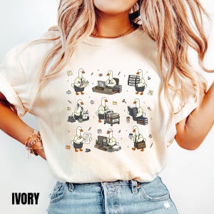 May include: An ivory t-shirt featuring a graphic print of nine cartoon geese dressed in office attire. The geese are depicted performing various legal tasks, including working at desks, carrying stacks of files, using telephones, and holding documents.