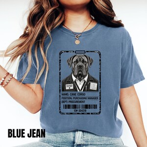 May include: A blue jean colored t-shirt featuring a graphic of an ID badge with a Cane Corso dog wearing a suit. The badge text reads: NAME: CANE CORSO, POSITION: PURCHASING MANAGER, DEPT: PROCUREMENT, ID# 004218, and PROCUREMENT SAVES MONEY.