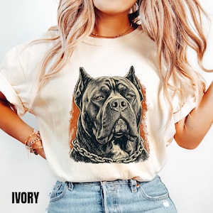 May include: An ivory short-sleeved t-shirt featuring a detailed black and white graphic illustration of a Cane Corso dog wearing a chain collar. The dog portrait is set against a textured, rectangular orange background on the front of the shirt.