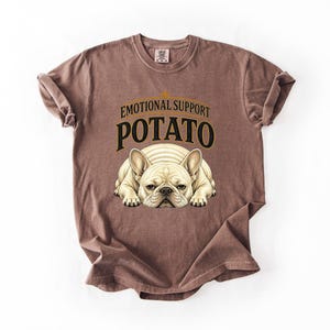 Könnte beinhalten: Ein braunes T-Shirt mit kurzen Ärmeln und einer Grafik eines cremefarbenen französischen Bulldoggen, der flach liegt. Über dem Hund steht der Text EMOTIONAL SUPPORT POTATO in schwarzen und goldenen Buchstaben. Das T-Shirt hat einen Rundhalsausschnitt.