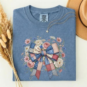 May include: A blue Comfort Colors sweatshirt featuring a floral bow design in red, white, and blue patchwork. The bow is surrounded by a variety of flowers in pink, white, and blue. A silver necklace with a star pendant is draped on the shirt.