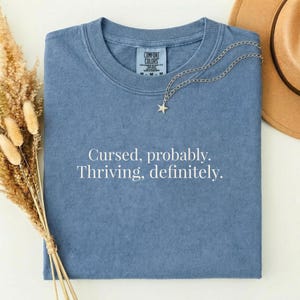 May include: A blue crew neck t-shirt with the white text Cursed, probably. Thriving, definitely. printed on the front. The shirt features a Comfort Colors brand label inside the collar and is accompanied by a silver star pendant necklace.