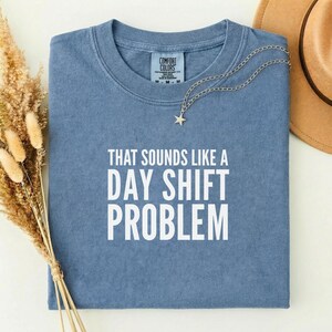 May include: A blue Comfort Colors t-shirt with the text "THAT SOUNDS LIKE A DAY SHIFT PROBLEM" in white. A silver necklace with a star pendant is draped over the shirt. A brown hat is visible in the background.