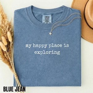 May include: A blue jean coloured t-shirt with a crew neckline and the white text "my happy place is exploring" printed on the front. The shirt features a Comfort Colors brand label at the inner neck and is accented by a silver star pendant necklace.