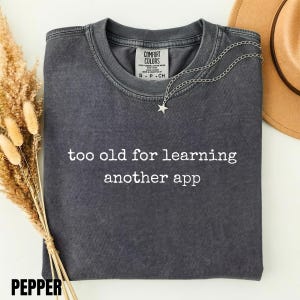 May include: A charcoal gray short-sleeve t-shirt with a crew neckline and the white text "too old for learning another app" printed on the front. The shirt features a Comfort Colors brand label inside the collar and a silver star necklace.