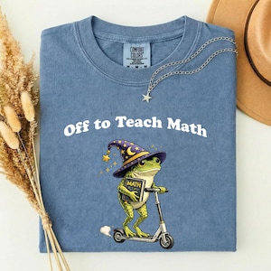 May include: Blue Comfort Colors t-shirt with the text "Off to Teach Math." The shirt features a cartoon frog wearing a wizard hat and riding a scooter while holding a book labelled "MATH."