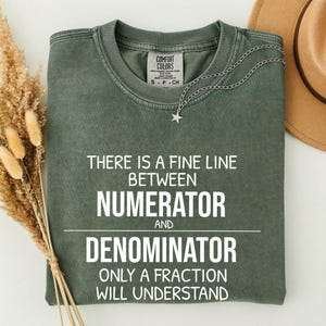 May include: Olive green t-shirt with the text "THERE IS A FINE LINE BETWEEN NUMERATOR AND DENOMINATOR ONLY A FRACTION WILL UNDERSTAND" in white. A silver necklace with a star pendant is draped across the shirt.