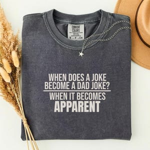 Peut inclure: T-shirt gris foncé avec le texte blanc : "WHEN DOES A JOKE BECOME A DAD JOKE? WHEN IT BECOMES APPARENT". Un collier argenté avec un pendentif étoile et un chapeau marron sont également visibles.