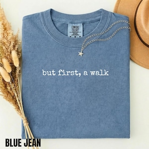 May include: A blue jean colored t-shirt with a crew neckline and short sleeves. The shirt features the white text but first, a walk printed across the chest. A silver star pendant necklace rests on the fabric, and the brand label reads Comfort Colors.