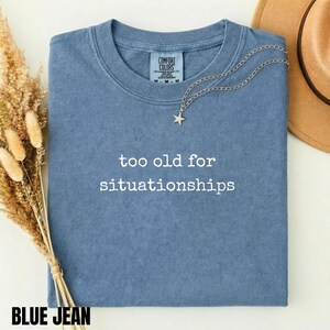 May include: A blue short-sleeved t-shirt with the white text "too old for situationships" printed on the front. The shirt features a crew neckline and a Comfort Colors brand label inside the collar. A silver star pendant necklace rests on the fabric.