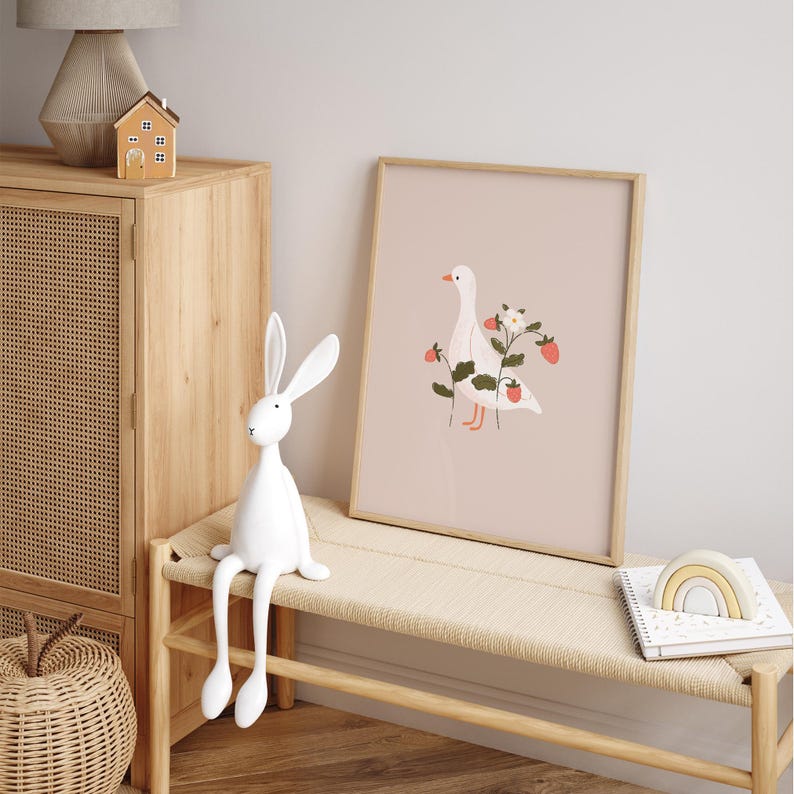 May include: A framed wall art print featuring a white goose illustration surrounded by green strawberry vines with red berries and a white flower on a solid light pink background. The artwork is displayed in a simple light wood frame.