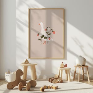 May include: A framed wall art print featuring a white goose standing among green strawberry plants with red berries and a white flower on a solid light beige background. The illustration is centered within a simple light wood frame.