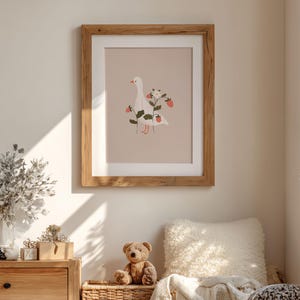 May include: A framed wall art print featuring a white goose illustration on a light beige background. The goose is surrounded by green stems, small white flowers, and red strawberries. The artwork is enclosed in a simple light wood frame.