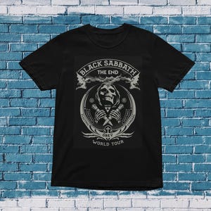 以下が含まれることがあります： ブラックサバスのグラフィックデザインが施された黒いTシャツ。デザインには、交差した楽器を持つスカル、バンド名、そして「THE END」という言葉が含まれています。「GREY WORLD TOUR」というテキストも含まれています。