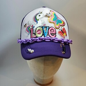 May include: A white and purple baseball cap with a decorative design. The cap features a unicorn, butterflies, and the word "LOVE" in colorful letters. A purple chain and pearls adorn the brim, with a black and gold charm on the side. The cap is displayed on a mannequin head.