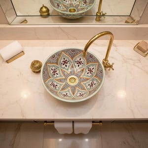 Op de afbeelding: Een decoratieve keramische wastafel met een bloemenpatroon in groen, rood en goud. De wastafel staat op een marmeren aanrecht met een gouden kraan en bijpassende hardware. Een spiegel weerspiegelt het ontwerp van de wastafel.