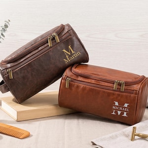 May include: Two brown leather toiletry bags with gold zippers. One bag has "M Martin" printed on it, and the other has "MICHAEL" printed on it. The bags are on a wooden surface.