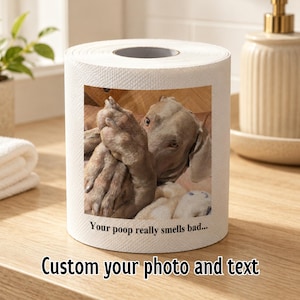 以下が含まれることがあります： 白いトイレットペーパーのロールで、犬の写真がプリントされています。犬は薄茶色で、ぬいぐるみと抱き合っているようです。ロールには「Your poop really smells bad...」と「Custom your photo and text」と書かれています。
