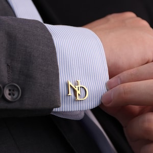 May include: Gold-colored cufflinks with the initials "ND" are displayed on a striped blue and white dress shirt. The cufflinks are attached to a gray suit jacket. The image is a close-up of the cufflink.