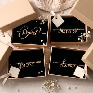 Puede incluir: Cuatro pulseras doradas con nombres, cada una exhibida en una pequeña caja de cartón abierta con interior negro. Los nombres "Daniel", "Marcus", "Janice" y "Louis" están escritos en cursiva. Cada caja tiene una pequeña etiqueta y está decorada con perlas y flores blancas.