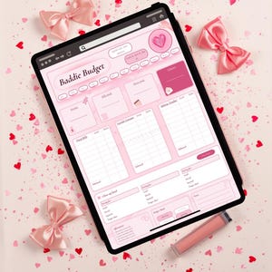 May include: A digital tablet displaying a pink-themed Baddie Budget financial planner interface with sections for income, bills, expenses, and savings goals, surrounded by pink satin bows and a lip gloss tube on a surface with scattered red heart confetti.