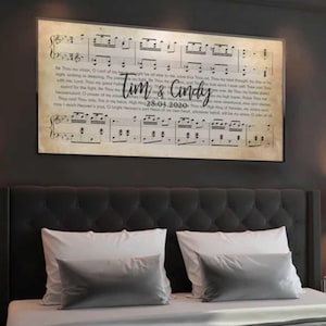 May include: A framed print featuring sheet music with the names "Tim & Cindy" and the date "28.03.2020" in a decorative font. The print is displayed above a bed with white pillows and a gray headboard.