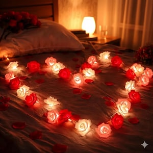 May include: A romantic scene featuring a heart-shaped arrangement of red and white rose string lights on a bed. Scattered rose petals add to the ambiance. A lamp and roses are visible in the background.