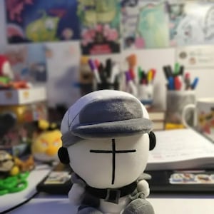 May include: A plush doll with a white body, gray hat, and black cross design on the face. The doll has a gray belt and is sitting on a white surface. The background is blurred, showing other objects.