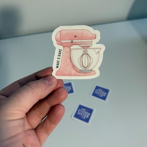May include: A pink sticker depicting a stand mixer with a whisk. The sticker has the text "WAKE & BAKE". Several smaller blue and white stickers are also visible.