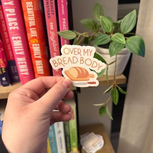 Könnte beinhalten: Ein Aufkleber mit dem Text "OVER MY BREAD BODY" und einer Cartoon-Illustration von Brot und einem Messer. Der Aufkleber wird vor einem Bücherregal mit Büchern und einer Topfpflanze gehalten.
