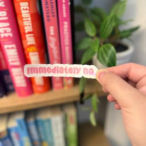 Puede incluir: Una pegatina rectangular rosa con el texto "immediately no.". Se sostiene frente a una estantería con lomos de libros coloridos. Los libros tienen títulos como "Eat Big Beautiful Life" y "Practice".